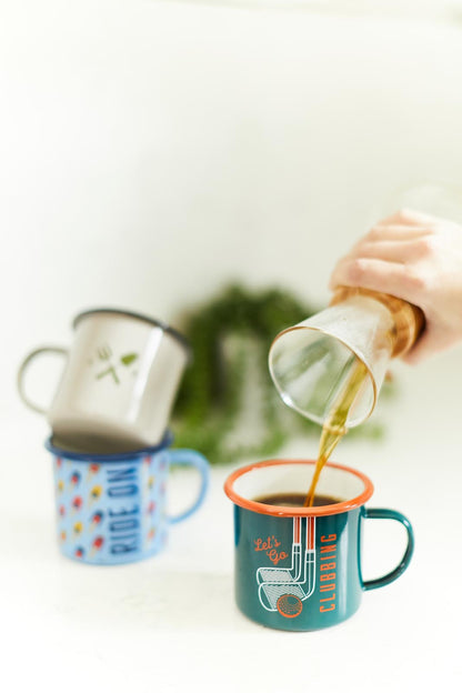 Enamel Mug - Cycling in green with orange rim, being filled with hot drink, with other patterned mugs in background.