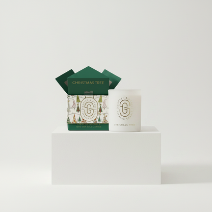 A white glass jar candle with a gold monogram logo sits next to its green and white illustrated gift box on a white pedestal against a light gray background.
