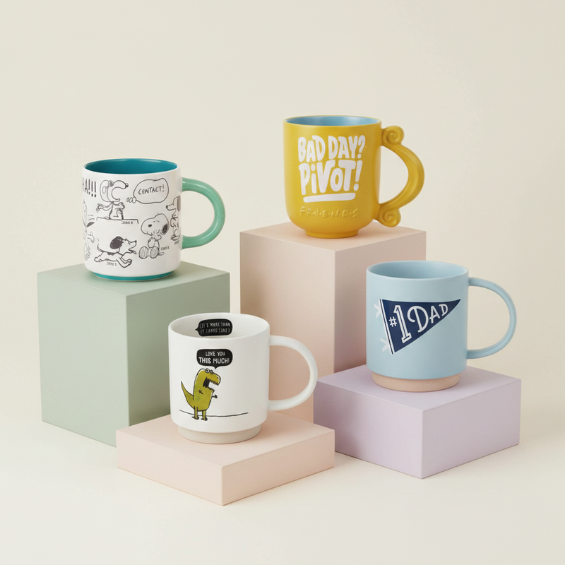 Four ceramic mugs displayed on pastel display blocks against a warm off-white background. The mugs include: a white mug with black Snoopy illustrations and a teal handle/interior; a yellow mug with a teal handle and interior reading 'BAD DAYS? PIVOT!'; a white mug with a yellow dinosaur and black text; and a light blue mug with a navy '1 DAD' pennant design.