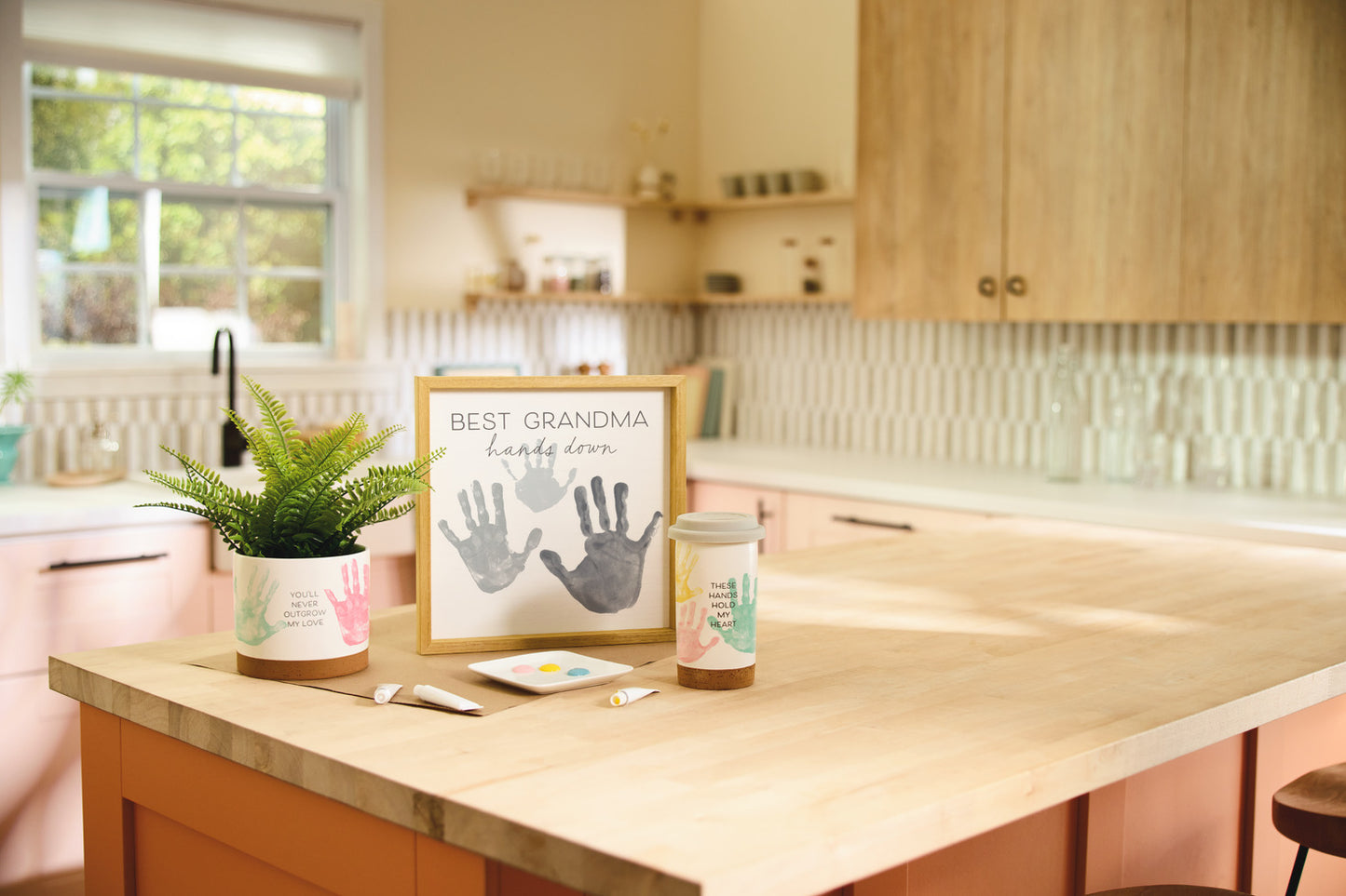 These Hands Hold My Heart Ceramic Travel Mug, 12.5 oz. on a kitchen table with a leafy plant and a framed handprint art.