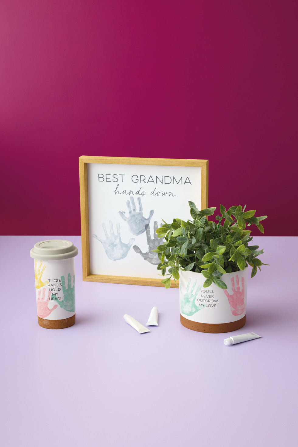These Hands Hold My Heart Ceramic Travel Mug, 12.5 oz. set on a purple table with a framed plaque and a plant in a pot.