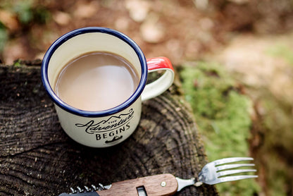 Enamel mug - Adventure Begins cream 11fl.oz/325ml showcasing warm beverage on a log with a camp fork, ideal for outdoor adventures.