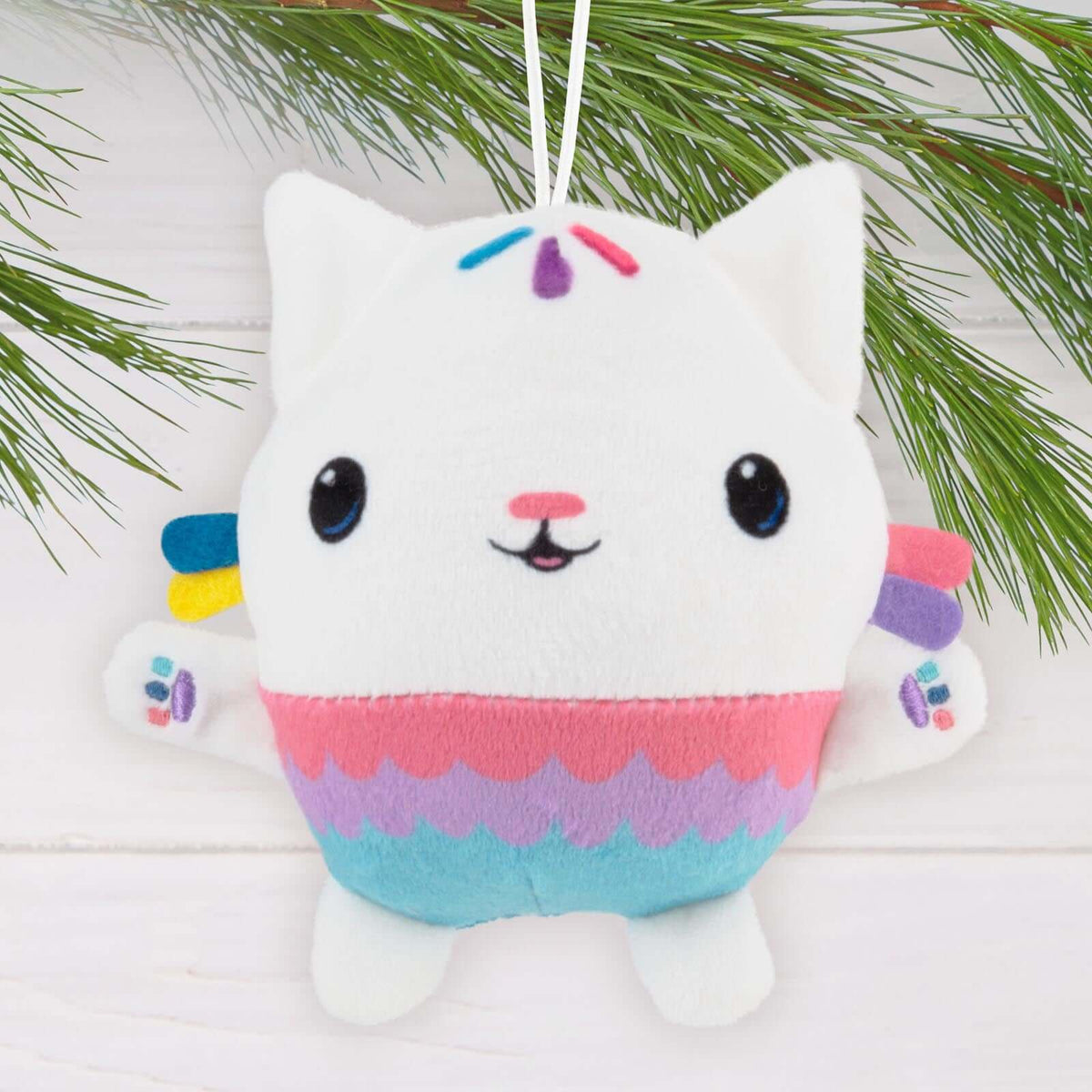 Gabby's Dollhouse Cakey Cat, Fabric - Hallmark Christmas Ornament, soft plush in pink and blue with a cheerful expression, hanging from a pine branch.