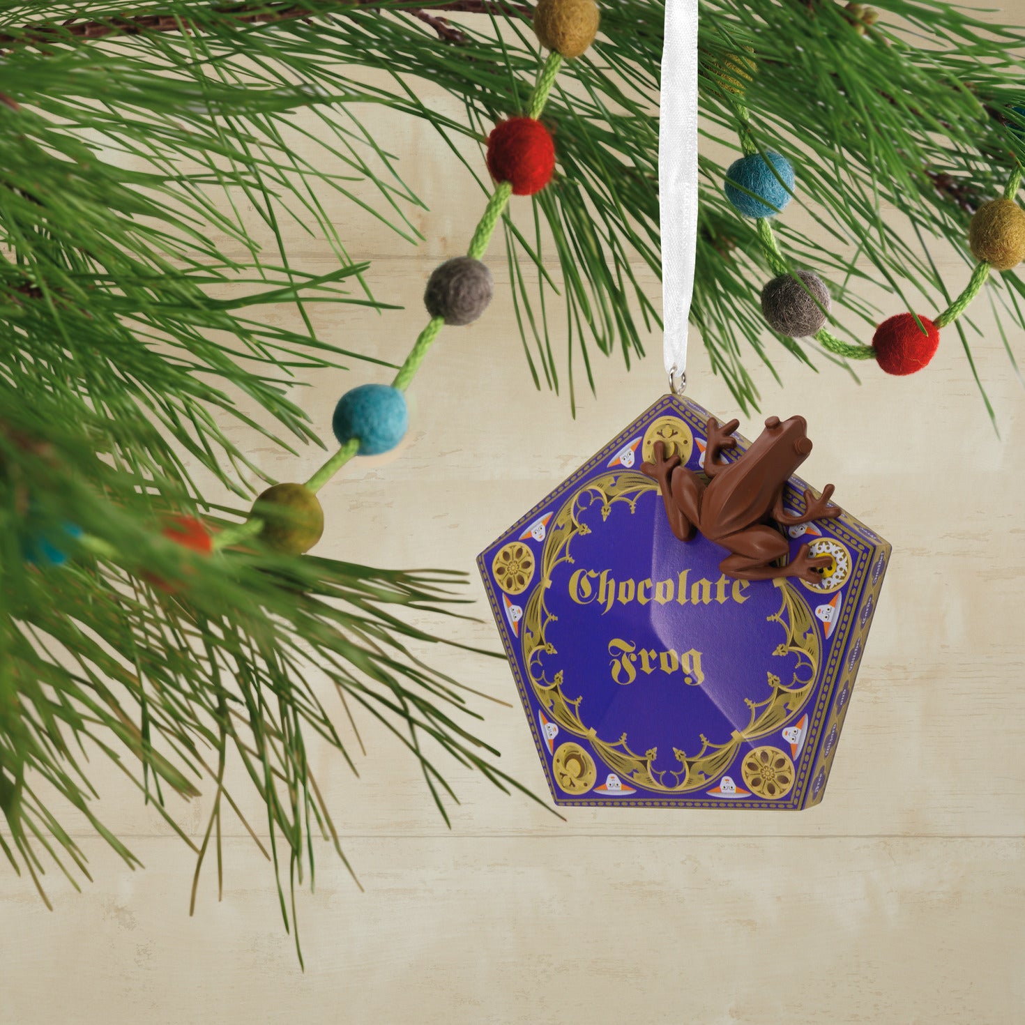 Hallmark Christmas Ornament - Harry Potter Chocolate Frog, hexagonal ornament design, featuring a chocolate frog perched on a purple candy box, dangling from festive greenery.