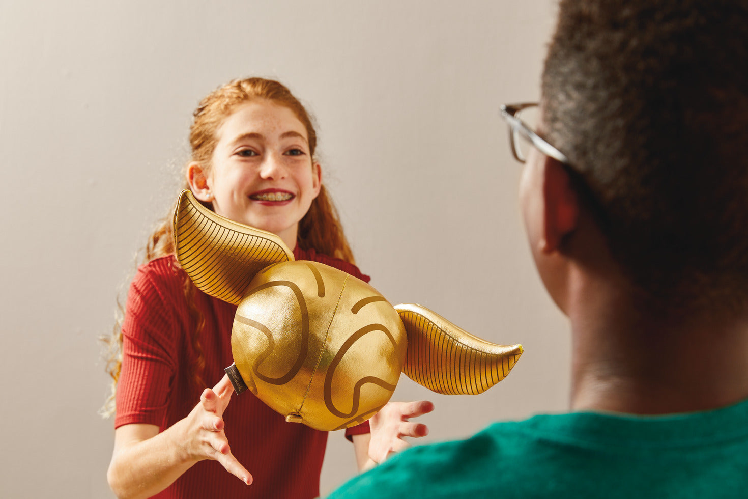 Harry Potter™ Golden Snitch™ Musical Plush With Motion, 8" being tossed by a girl in a red top to a boy in a green shirt.