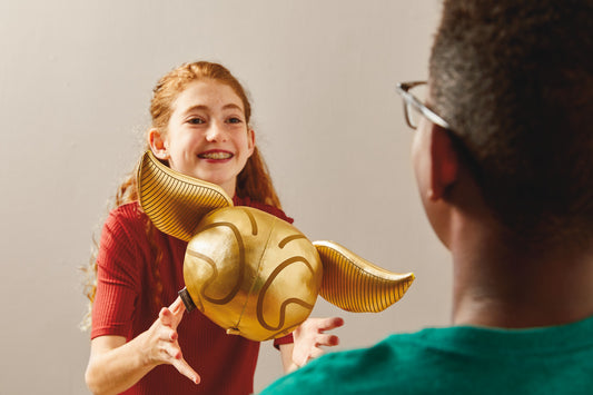 Harry Potter™ Golden Snitch™ Musical Plush With Motion, 8" being tossed by a girl in a red top to a boy in a green shirt.