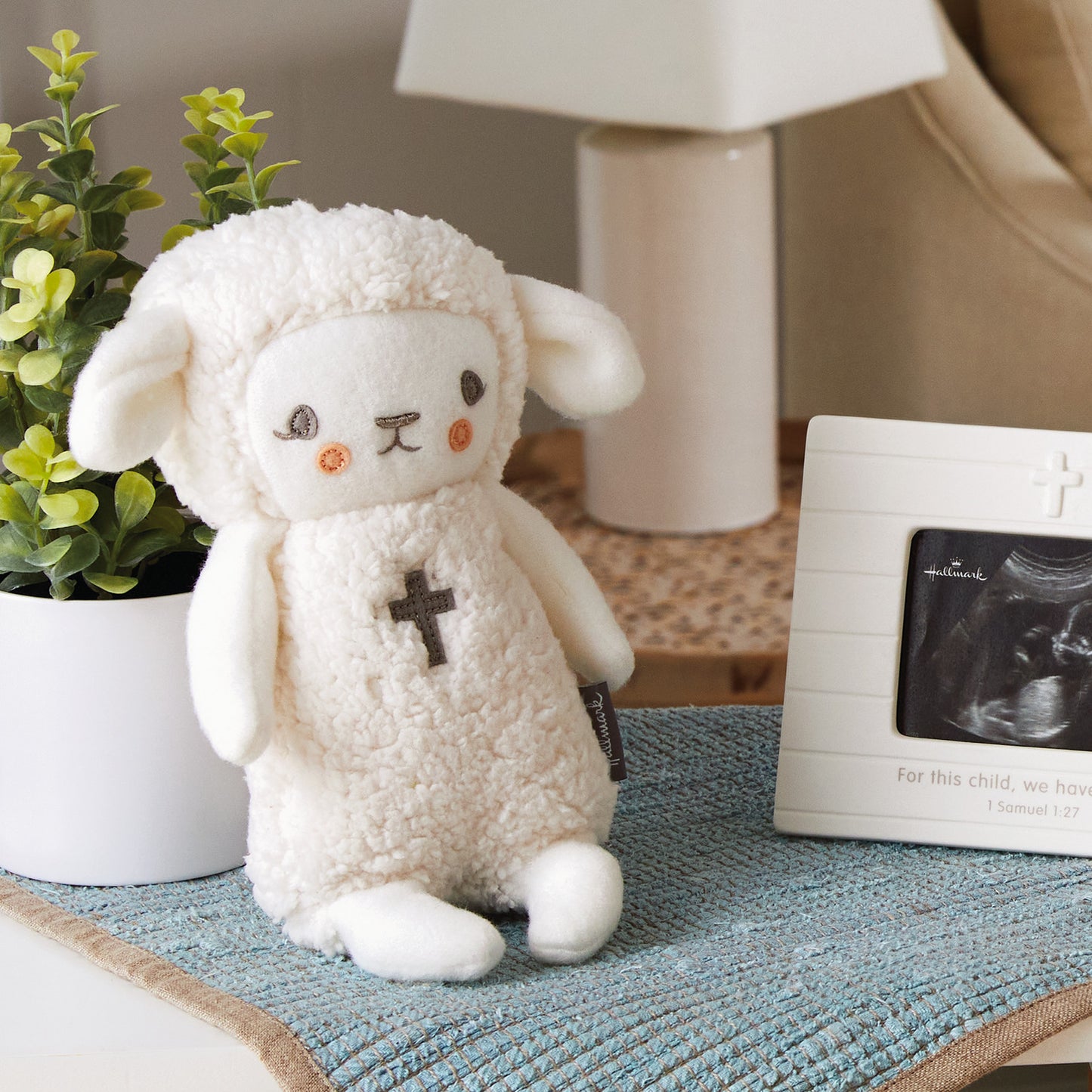 Lullaby Lamb Musical Stuffed Animal, 8.25", soft plush toy with embroidered cross, displayed on a table with greenery.