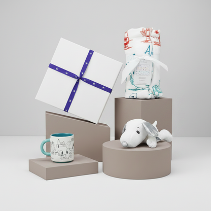 A styled gift set featuring a white gift box with a purple ribbon, a white baby blanket with red and teal illustrations wrapped in a white ribbon, a white and teal patterned mug, and a white plush toy, all arranged on matte taupe pedestals against a light gray background.