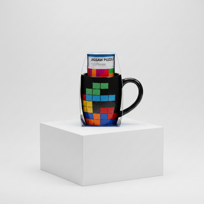 A black ceramic mug with a colorful Tetris puzzle design, shown with its clear plastic packaging standing inside it on a white display block against a light gray studio background.