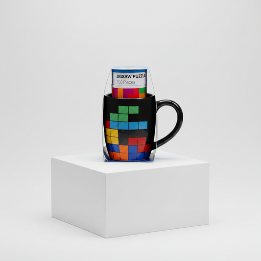 A black ceramic mug with a colorful Tetris puzzle design, shown with its clear plastic packaging standing inside it on a white display block against a light gray studio background.