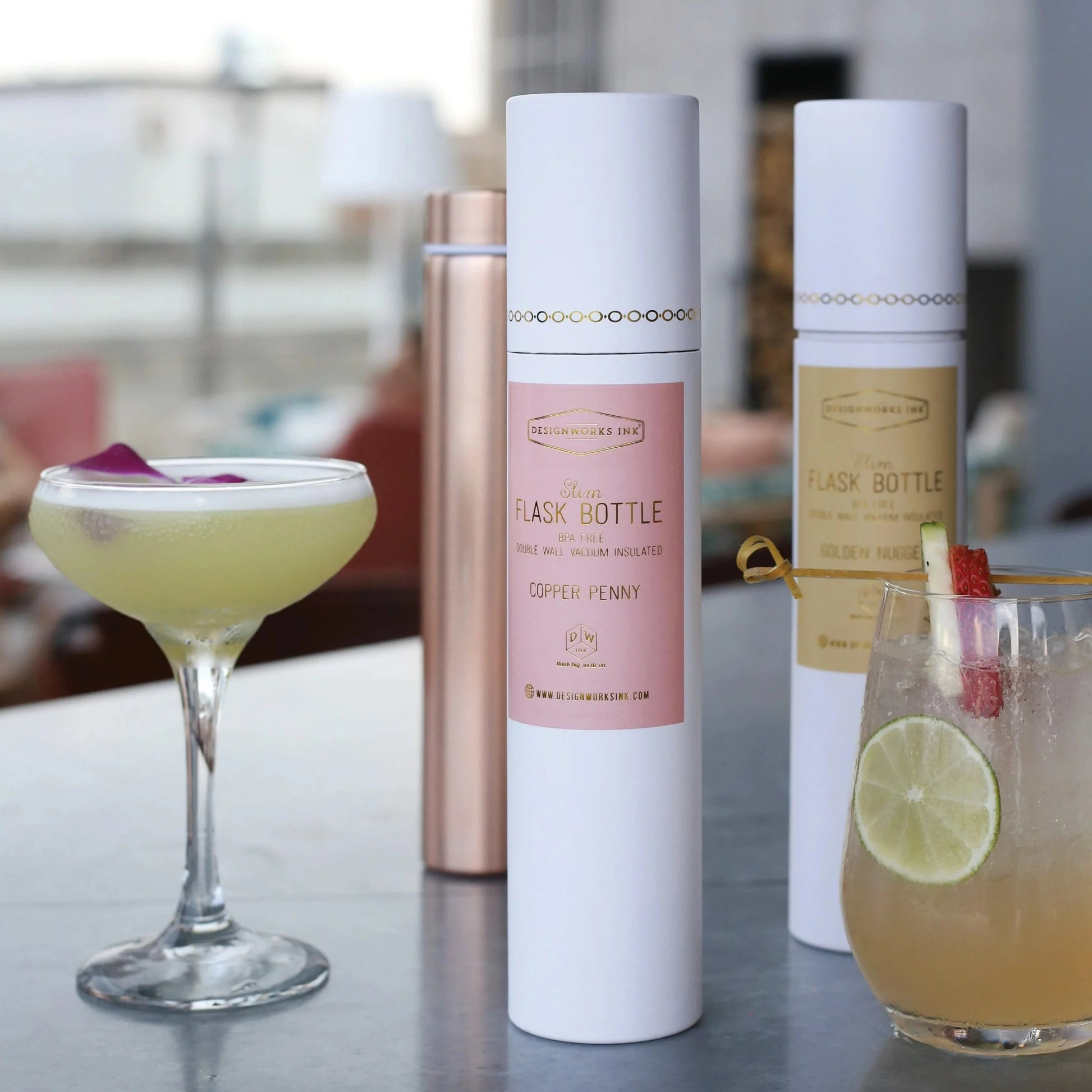 White slim flask bottle in a foil-stamped tube, displayed alongside cocktails and other flask bottles, featuring a sleek, modern design.